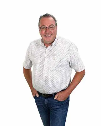 Man met een wit-blauw gestipte blouse en een bril, met zijn handen in zijn zakken.