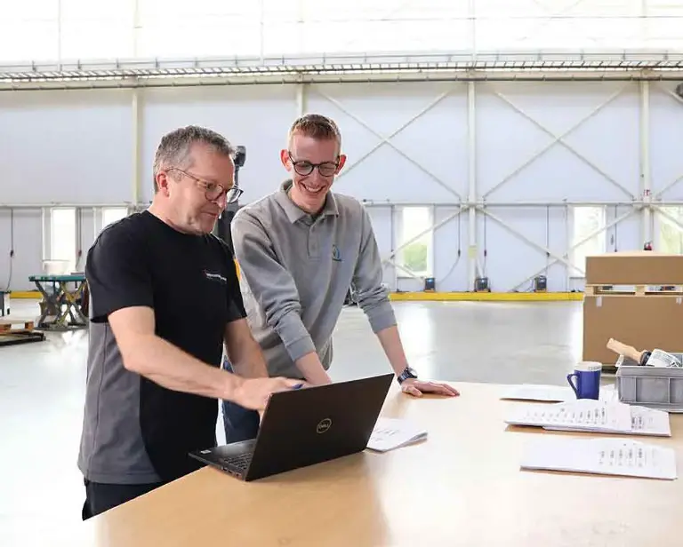 Twee personen staan aan een tafel en kijken naar een laptop.