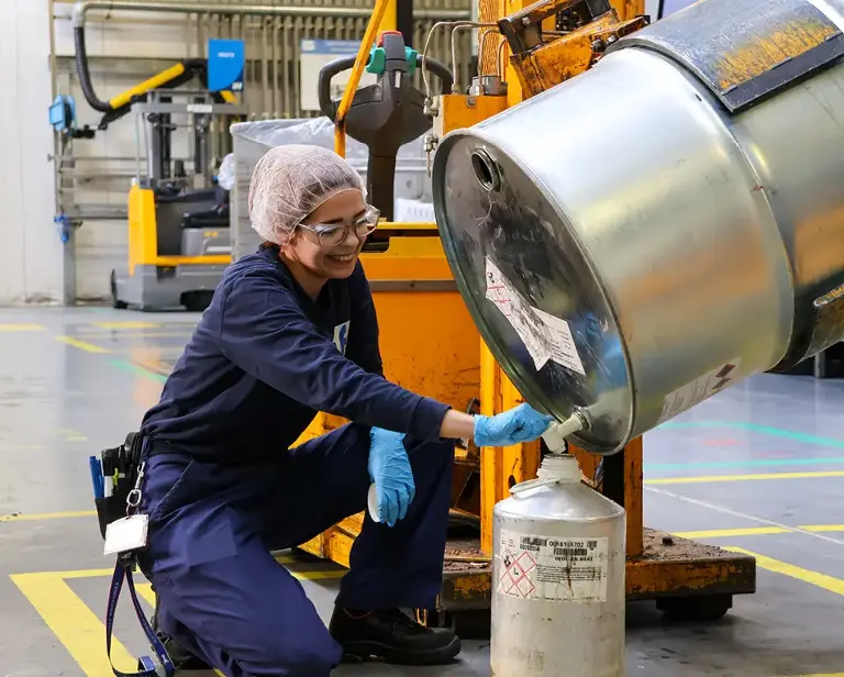 Persoon in een fabriek met blauwe overal en haar netje giet vloeistof uit een grote metalen ton in een kleiner vat.