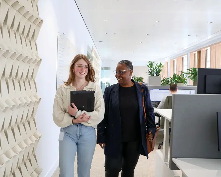 Twee personen lopen door een kantoorruimte, een draagt een laptop, de ander een tas.