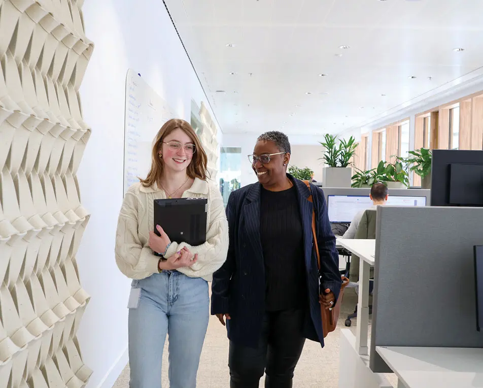 Twee personen lopen door een kantoorruimte, een draagt een laptop, de ander een tas.