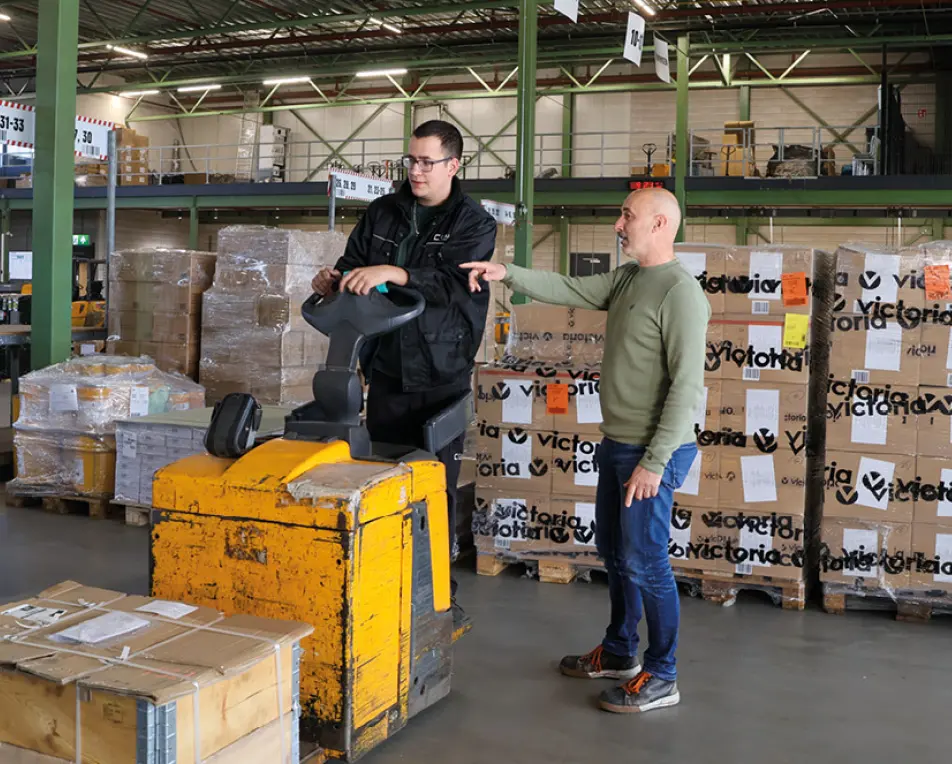 Twee personen in een magazijn; één bedient een gele palletwagen terwijl de ander wijst ergens naartoe.