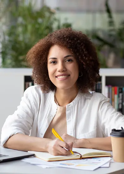 Vrouw met krullen zit in een kantoor en schrijft in een notitieboek.