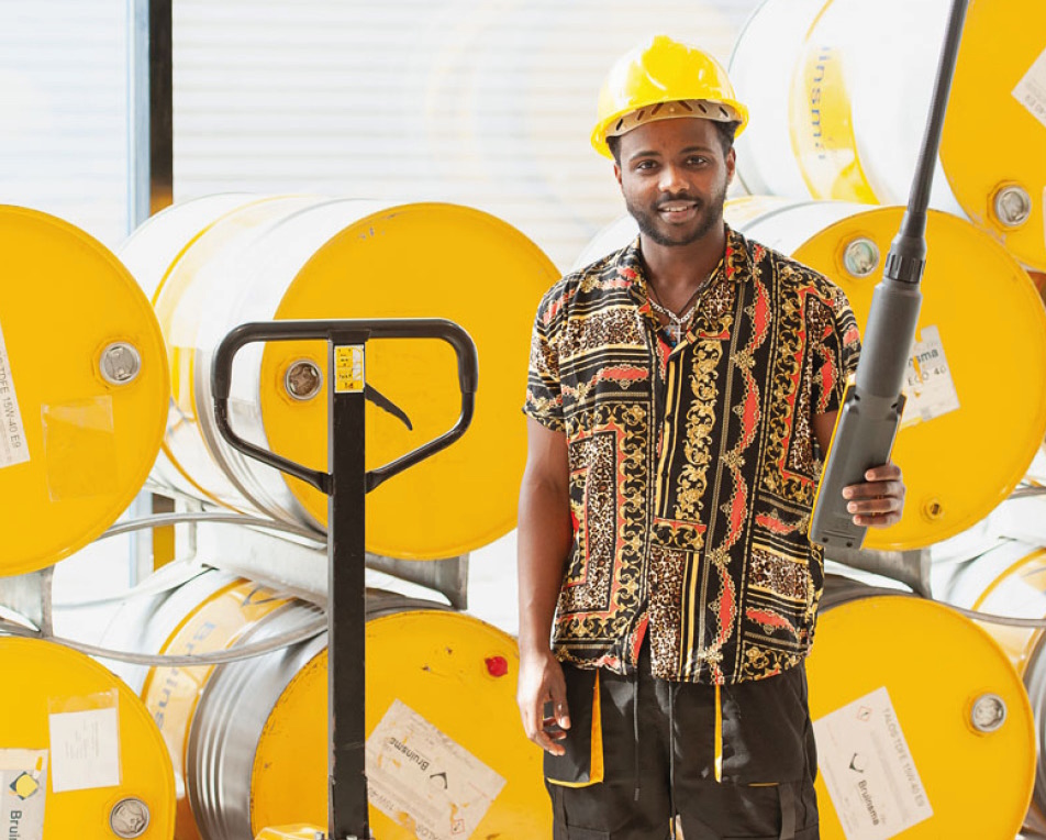 Man in werkkleding en veiligheidshelm, met gele tonnen in de achtergrond.