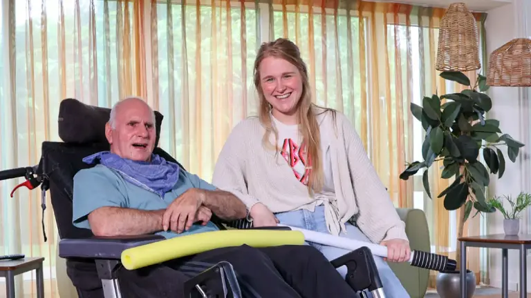 Man in rolstoel en vrouw met beige trui met een woonkamer als achtergrond.