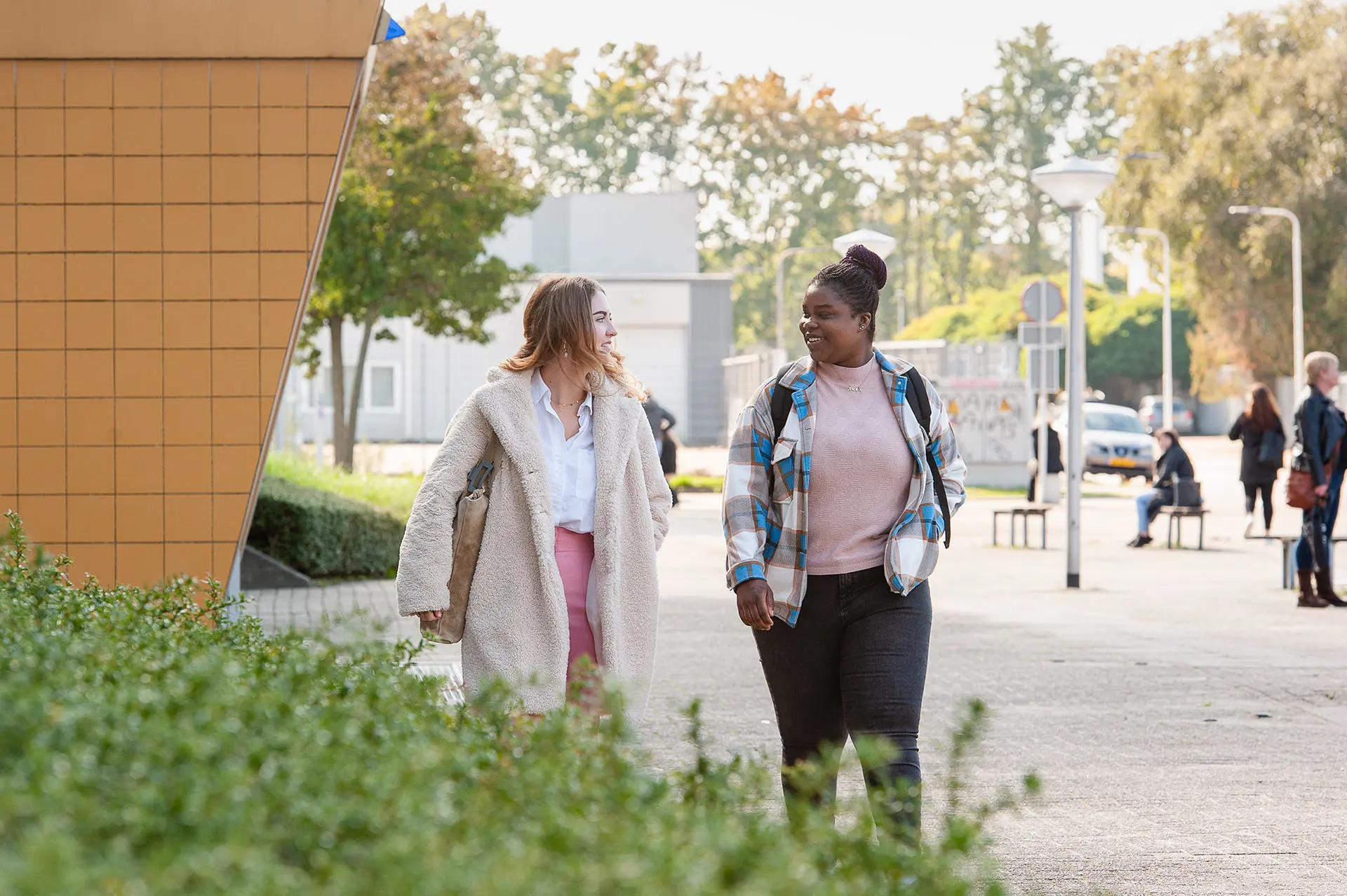Twee personen lopen over een plein. In de achtergrond staan bomen en andere mensen.
