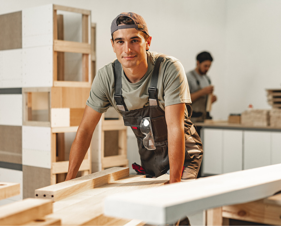 Twee personen in een houtbewerkingswerkplaats; de voorste draagt een pet, grijs T-shirt en overall met veiligheidsbril, leunend op een houten werkbank