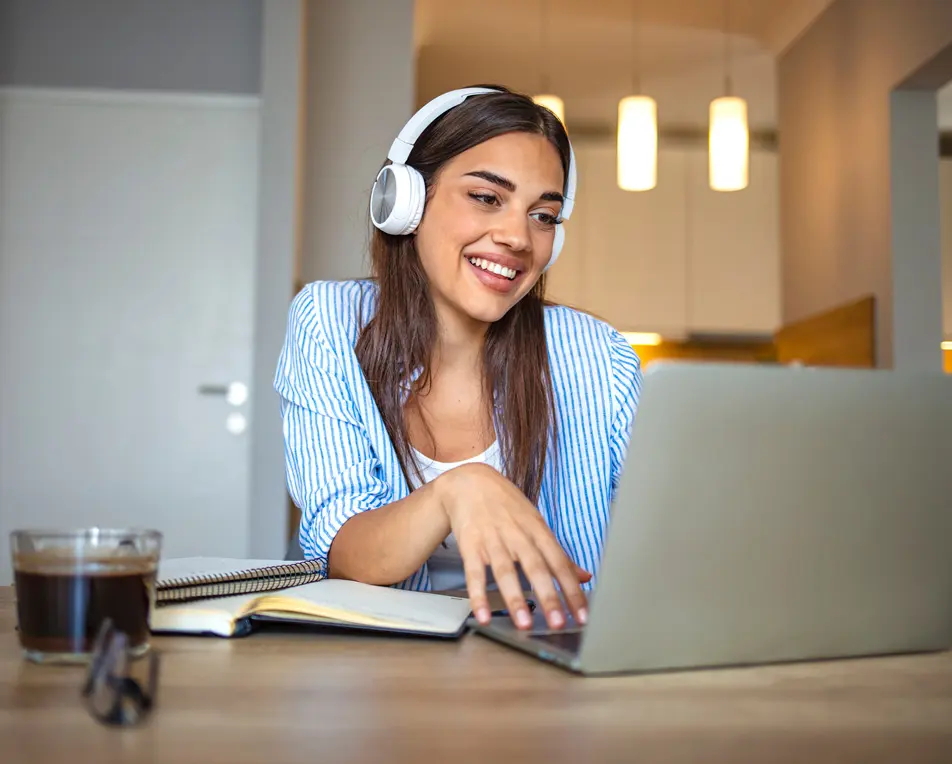 Persoon met koptelefoon zit aan een tafel met een laptop, notitieboek en een glas donkere drank, in een sfeervol verlichte binnenruimte