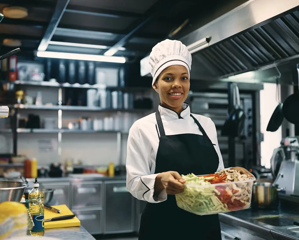 Chef-kok met witte koksmuts en zwart schort houdt een doorzichtige bak met gesneden groenten vast in een professionele keuken met roestvrijstalen apparatuur.