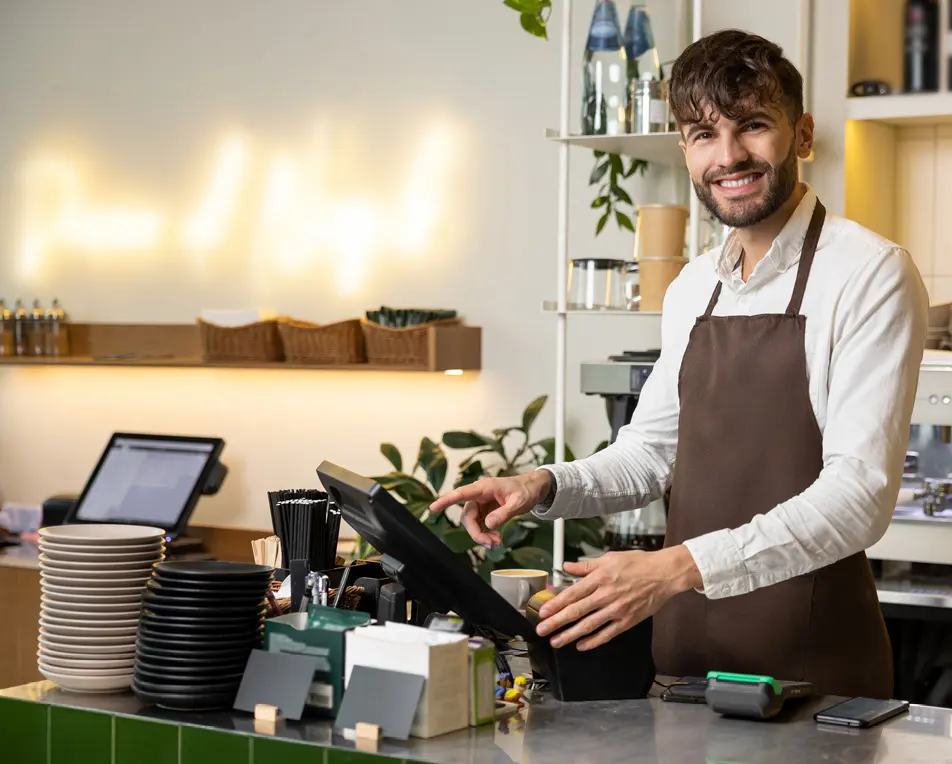 Barista met bruine schort bedient een touchscreen-kassa achter de toonbank van een koffiezaak, met kopjes, borden en een espressomachine op de achtergrond.