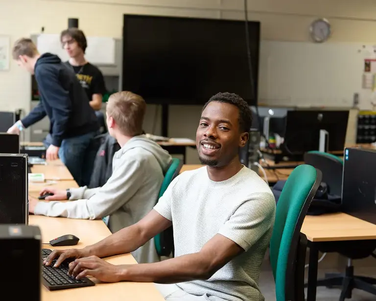 Computerlokaal met daarin personen, de voorste persoon heeft een grijs shirt aan en is aan het typen.