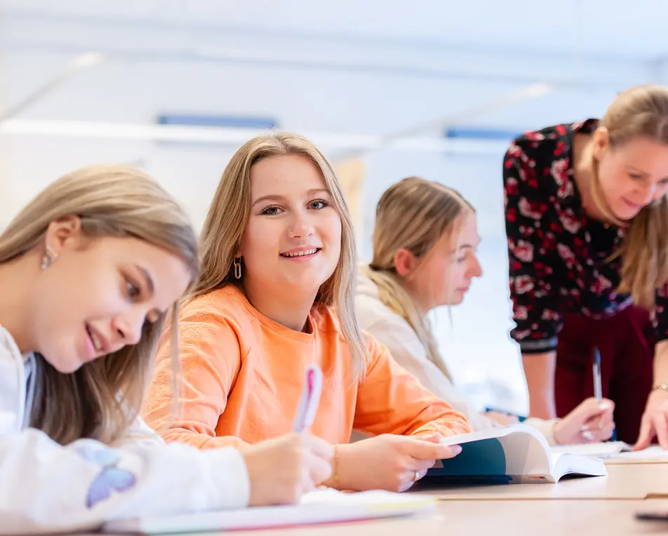Drie studenten zitten aan een tafel met boeken, terwijl een docent erbij staat in een klaslokaal.