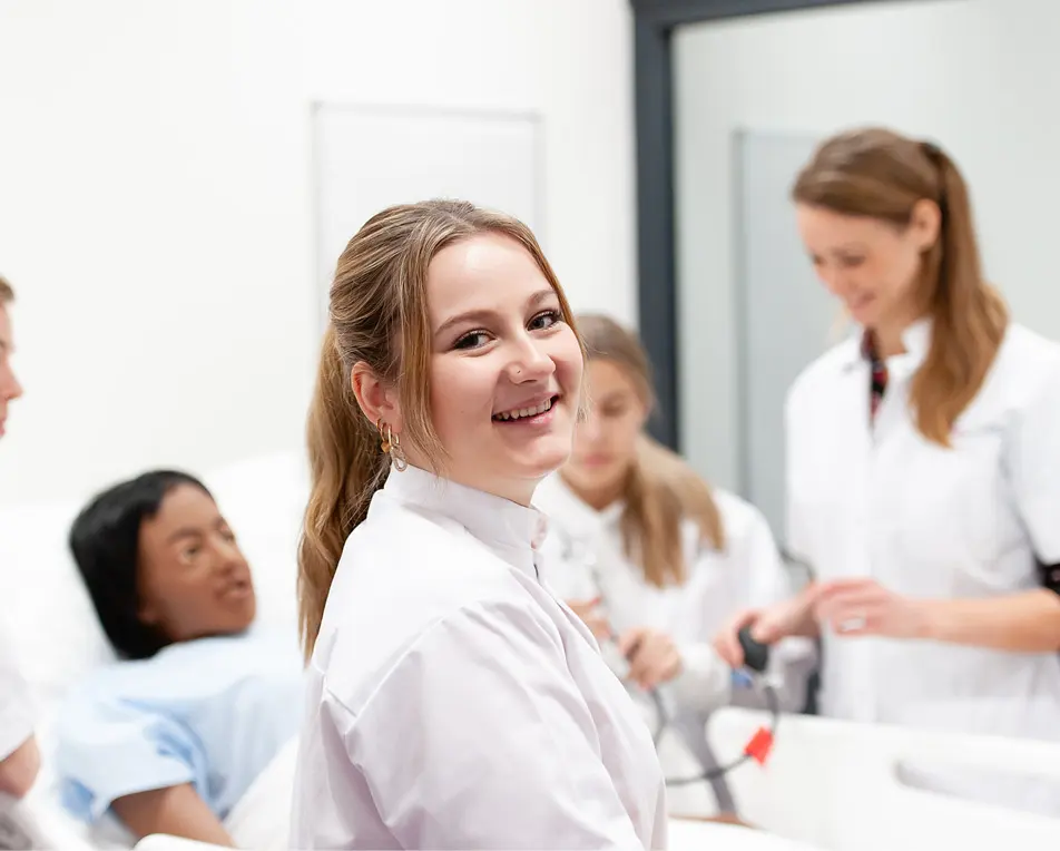 Groep mensen in witte jassen rond een bed in een medische of opleidingssetting.