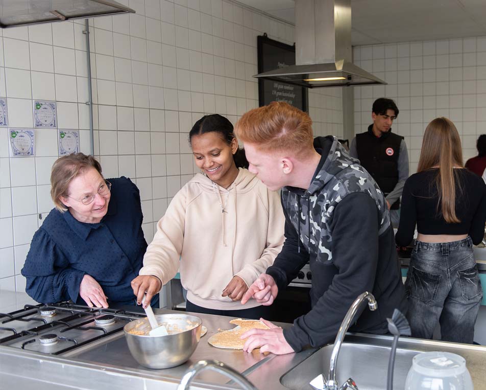 Een keuken met op de voorgrond drie personen. Links een oudere vrouw die kijkt naar een jonge jonge die rechts staat en in het midden een meisje die aan het roeren is.
