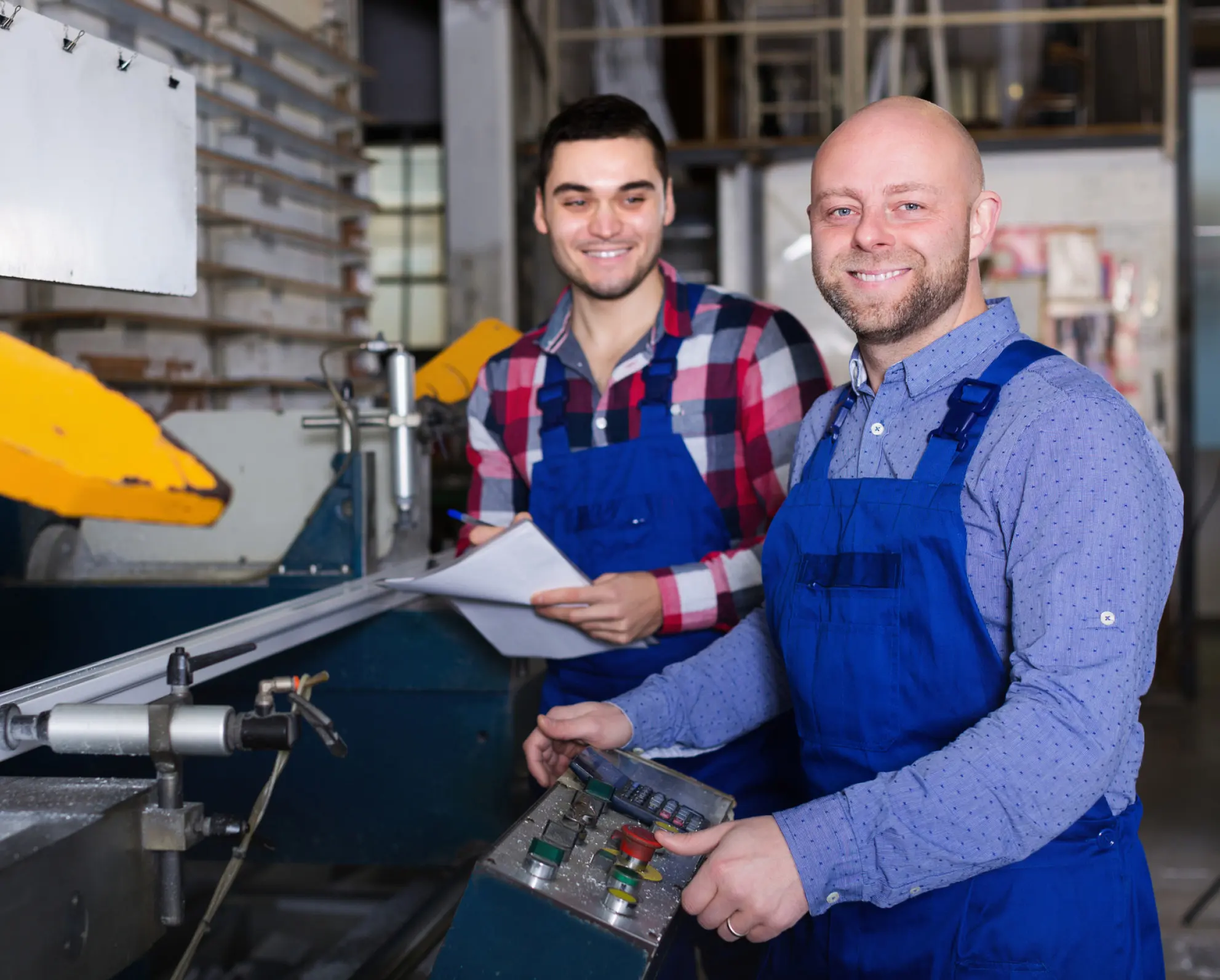 Twee personen in blauwe overalls werken bij machines in een industriële omgeving.