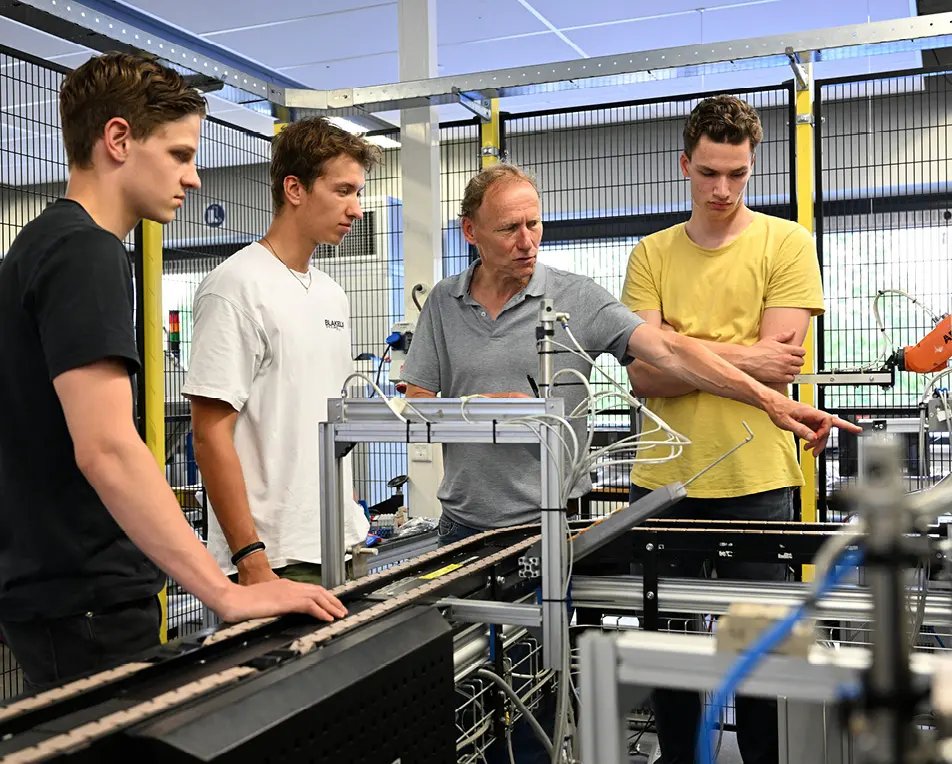 Vier personen staan rond industriële apparatuur in een technische of educatieve omgeving.
