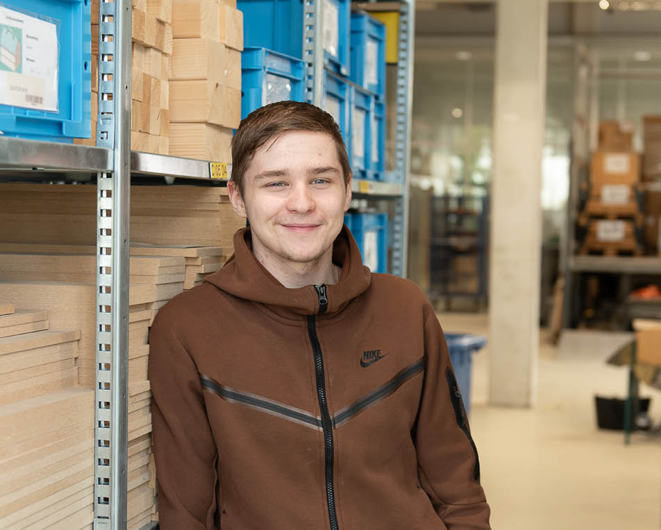 Persoon met een bruine Nike-hoodie staat in een opslagruimte met stellingen vol blauwe plastic bakken en stapels houten planken