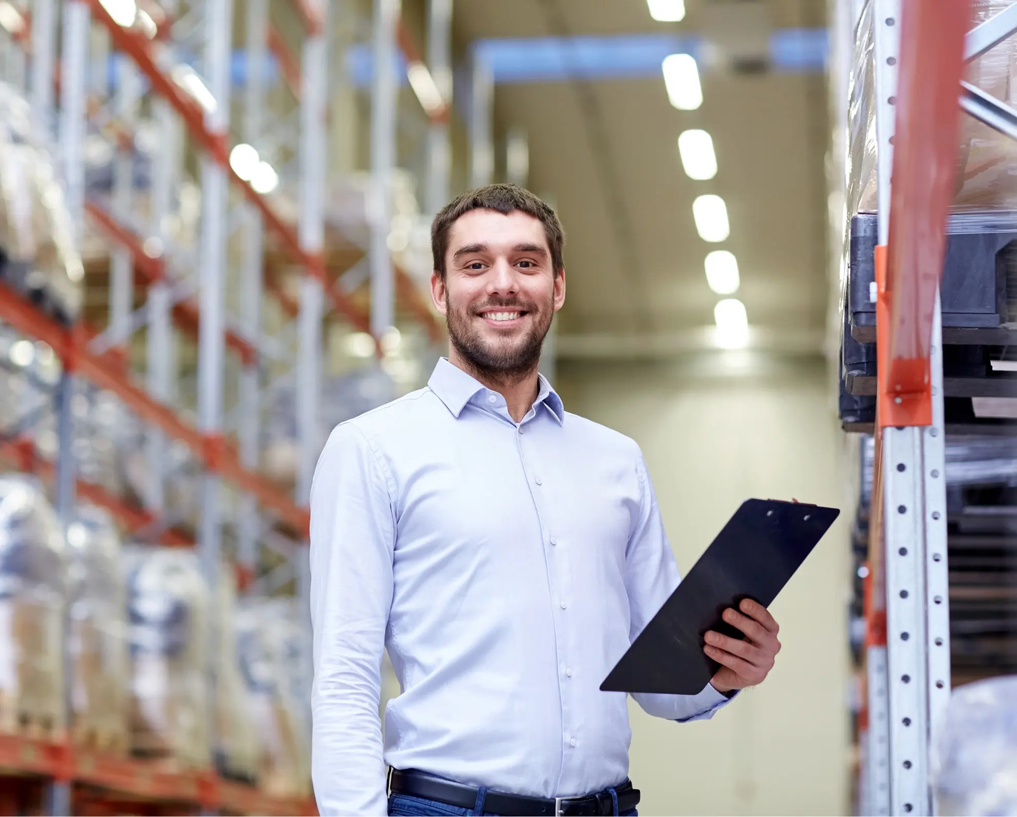Persoon met een lichtblauw overhemd en donkere broek staat in een magazijn en houdt een clipboard vast