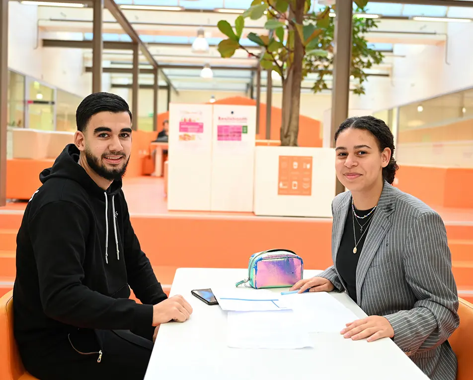 Twee personen zitten in een moderne, lichte ruimte met glazen wanden; één draagt een bruine jas en gescheurde jeans, de ander een beige trui en zit aan een tafel.