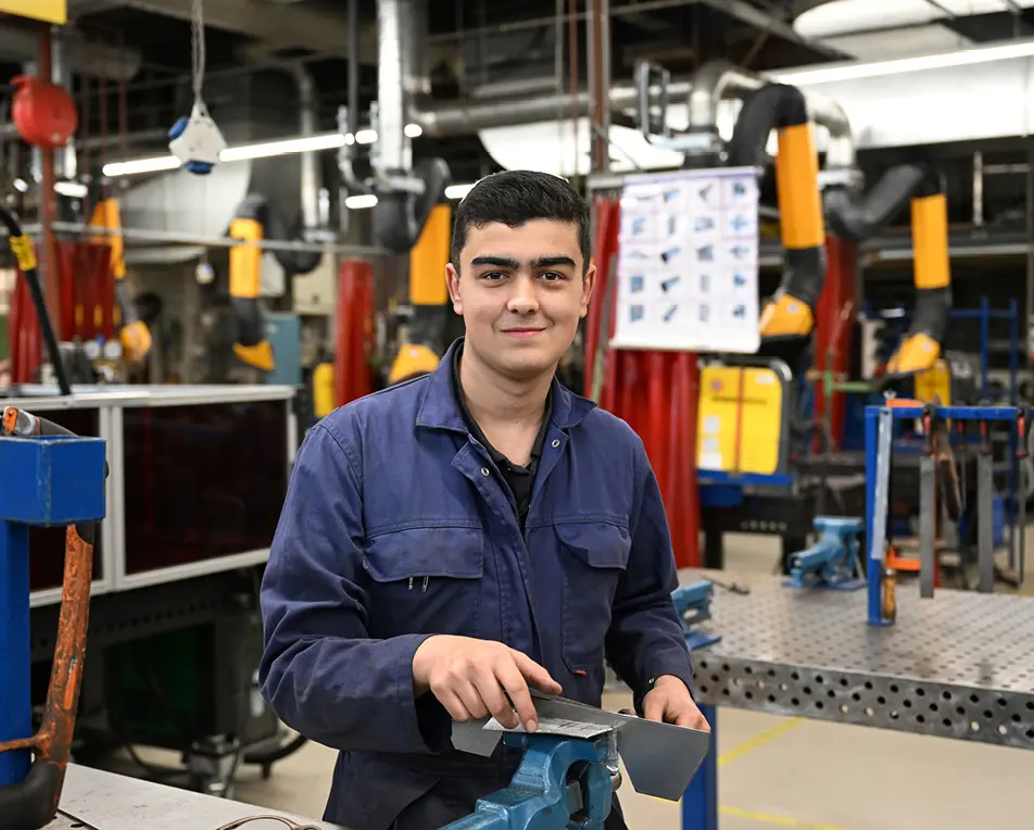 Persoon in blauwe werkjas werkt met een stuk metaal aan een werkbank in een industriële werkplaats.