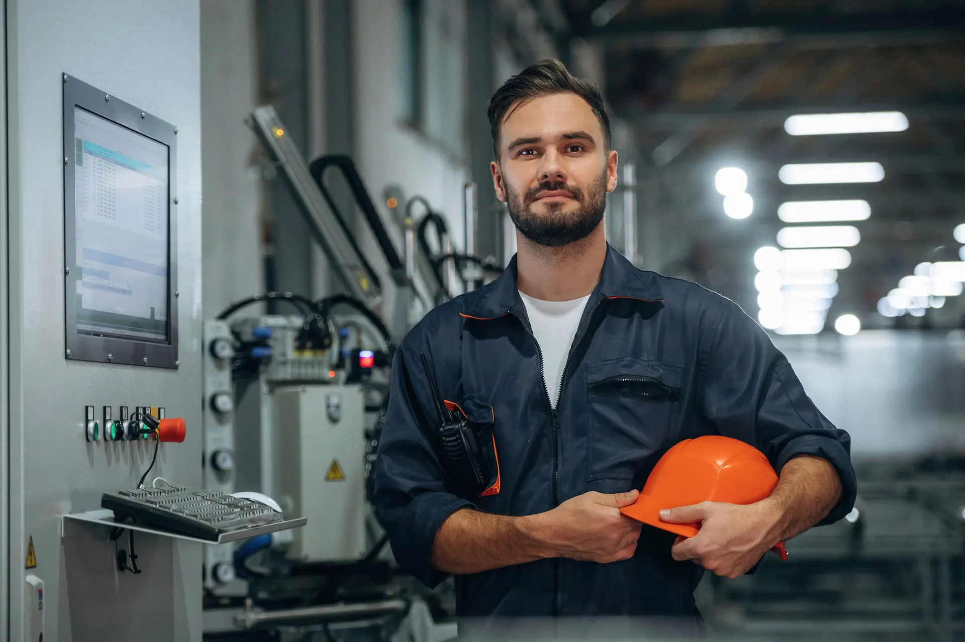 Persoon in werkuniform met oranje helm in een industriële omgeving met machines en een bedieningspaneel.