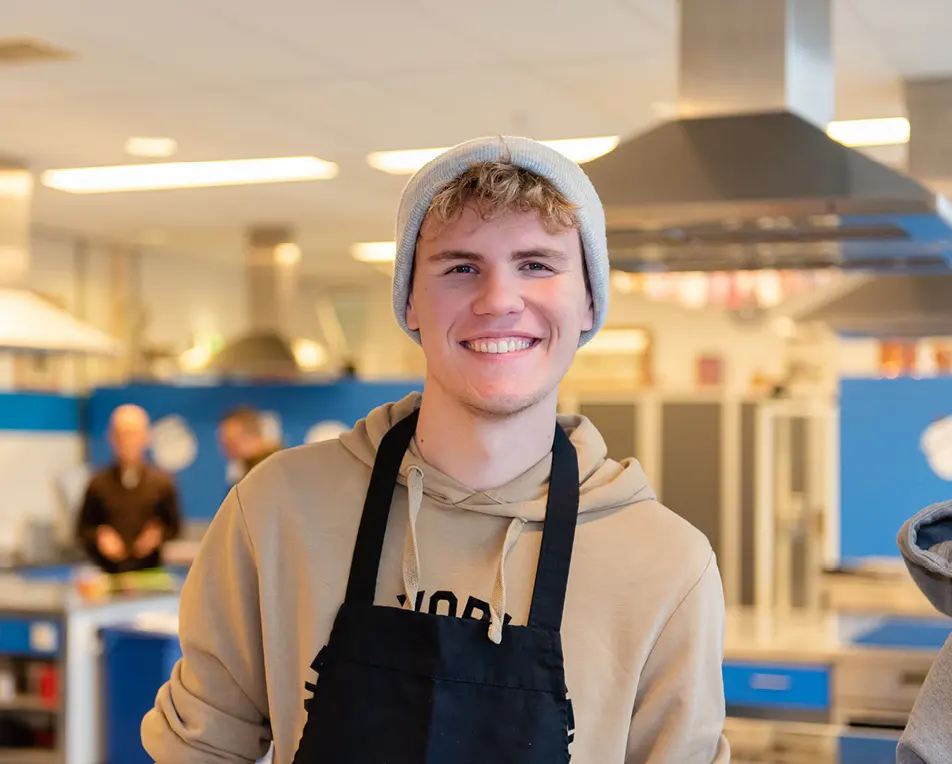 Persoon met een beige hoodie en een zwart schort staat in een keuken