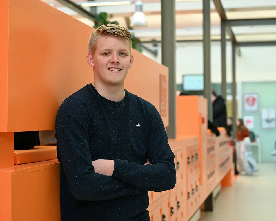 Persoon met donkere lange mouwen en gekruiste armen staat voor een rij oranje lockers in een binnenruimte met glazen plafond
