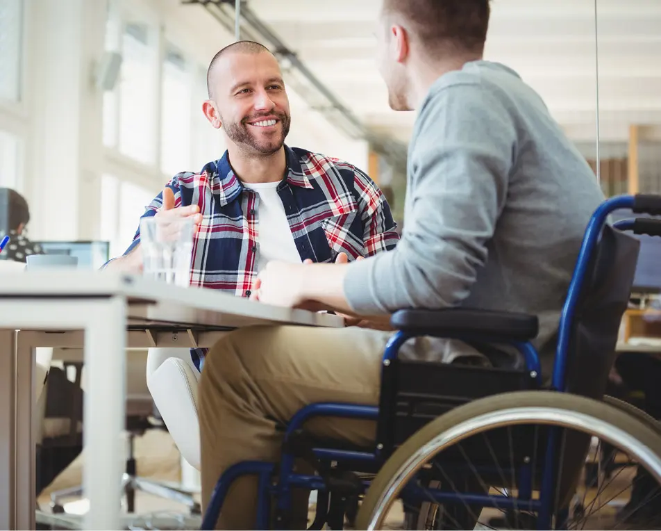 Twee personen zitten aan een tafel, een zit in een rolstoel.