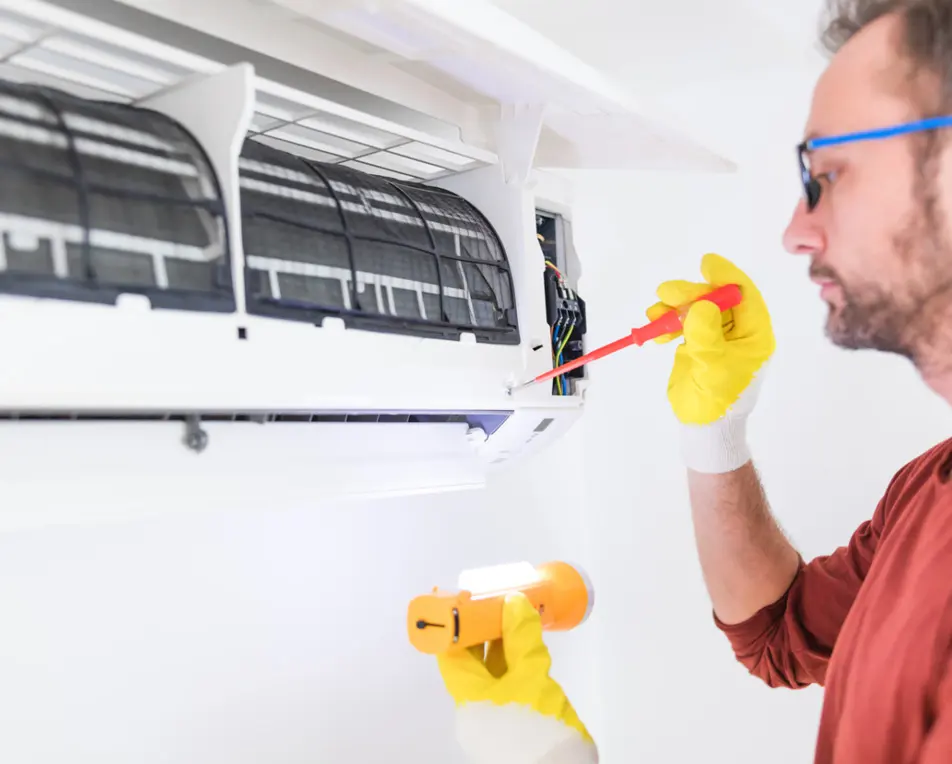 Een persoon voert onderhoud uit aan een airco met een schroevendraaier en een zaklamp, en draagt gele handschoenen.