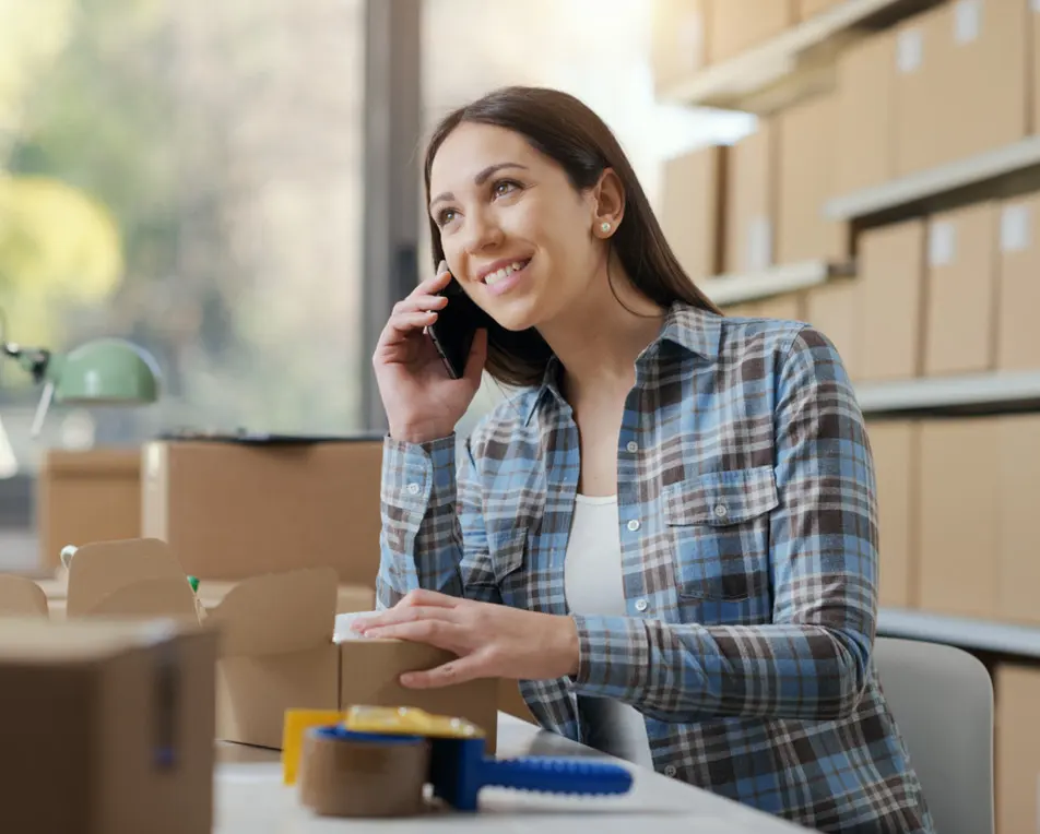 Vrouw in geruite blouse is aan het bellen, omringt door dozen.