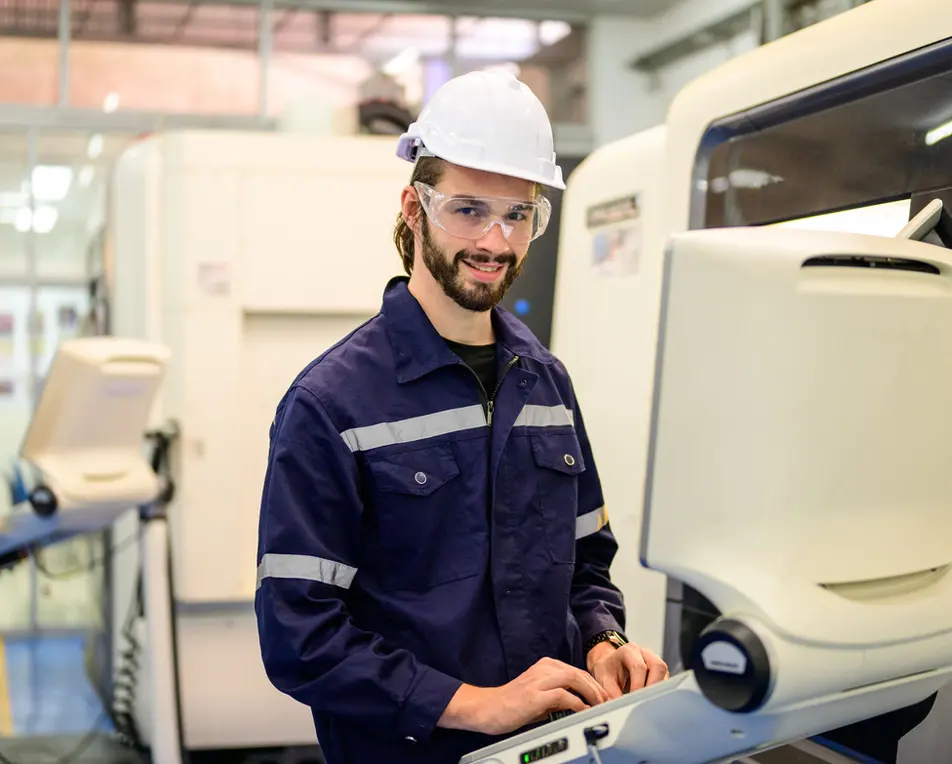 Persoon met witte veiligheidshelm en blauw werkuniform met reflecterende strepen werkt op een computer in een industriële omgeving met machines op de achtergrond