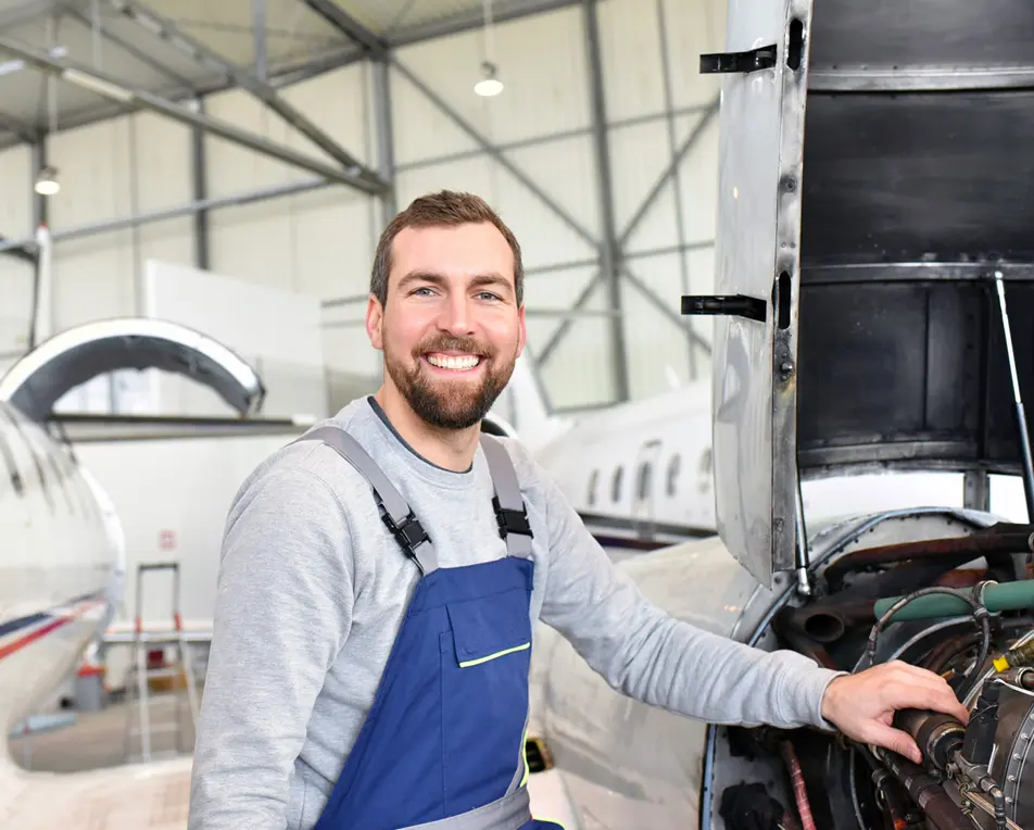 Persoon in een hangar werkt aan een vliegtuigmotor, gekleed in een grijze longsleeve en blauwe overall, met andere vliegtuigen zichtbaar op de achtergrond