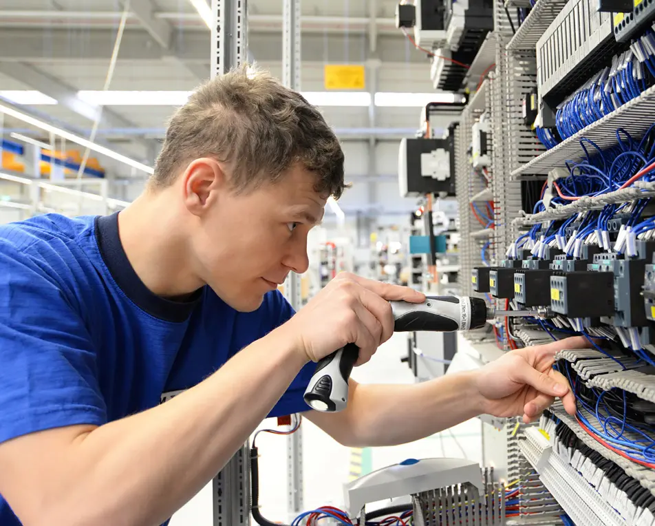 Persoon werkt met een schroevendraaier aan een elektrisch paneel vol bedrading in een technische omgeving.