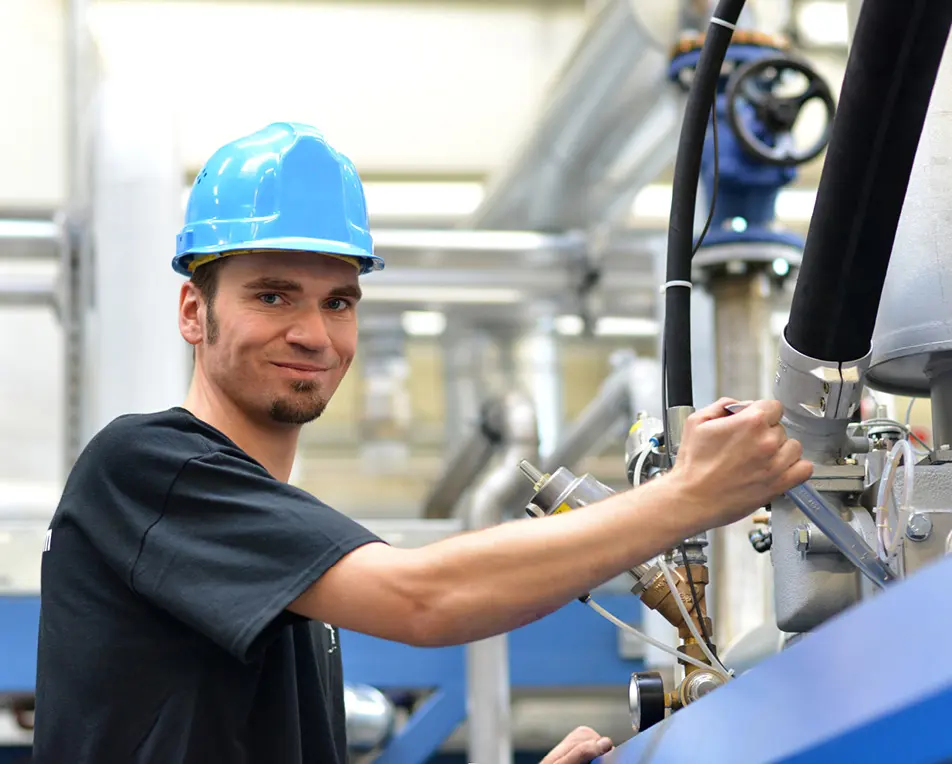 Een persoon met een blauwe veiligheidshelm en zwarte kleding bedient een machine in een industriële omgeving.