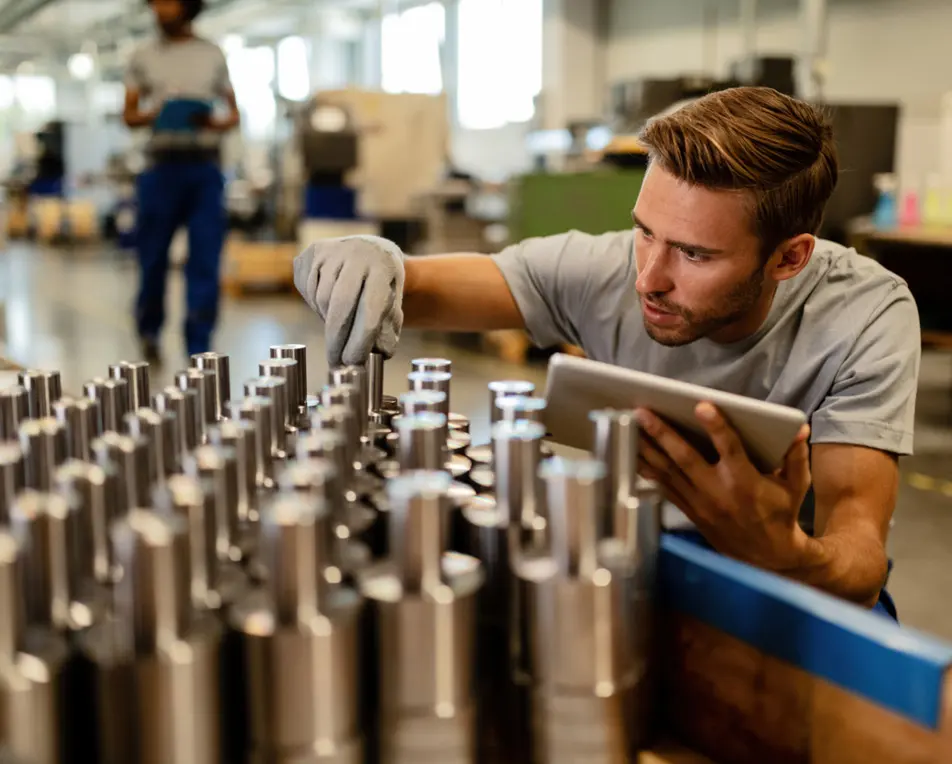 Persoon inspecteert metalen onderdelen met een tablet in een industriële omgeving; tweede persoon op de achtergrond.