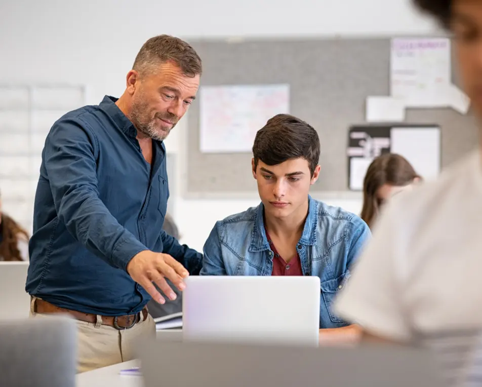 Twee personen zitten in een klaslokaal. Eén achter een laptop en één staat er langs.