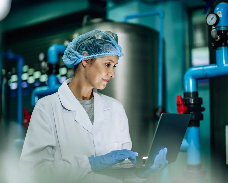 Persoon met witte labjas, blauwe handschoenen, haarnetje en veiligheidsbril werkt op een laptop in een industriële omgeving met blauwe leidingen en machines op de achtergrond.