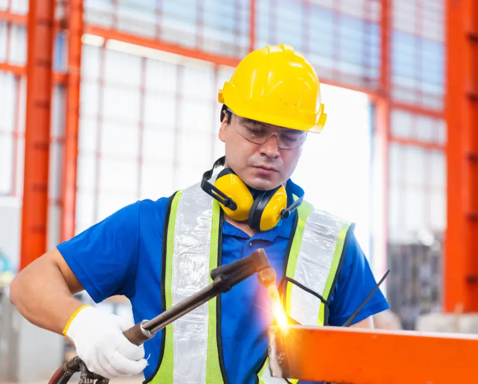 Bouwvakker met gele helm en lasapparaat aan het lassen bij grote oranje balken in een industriële omgeving.
