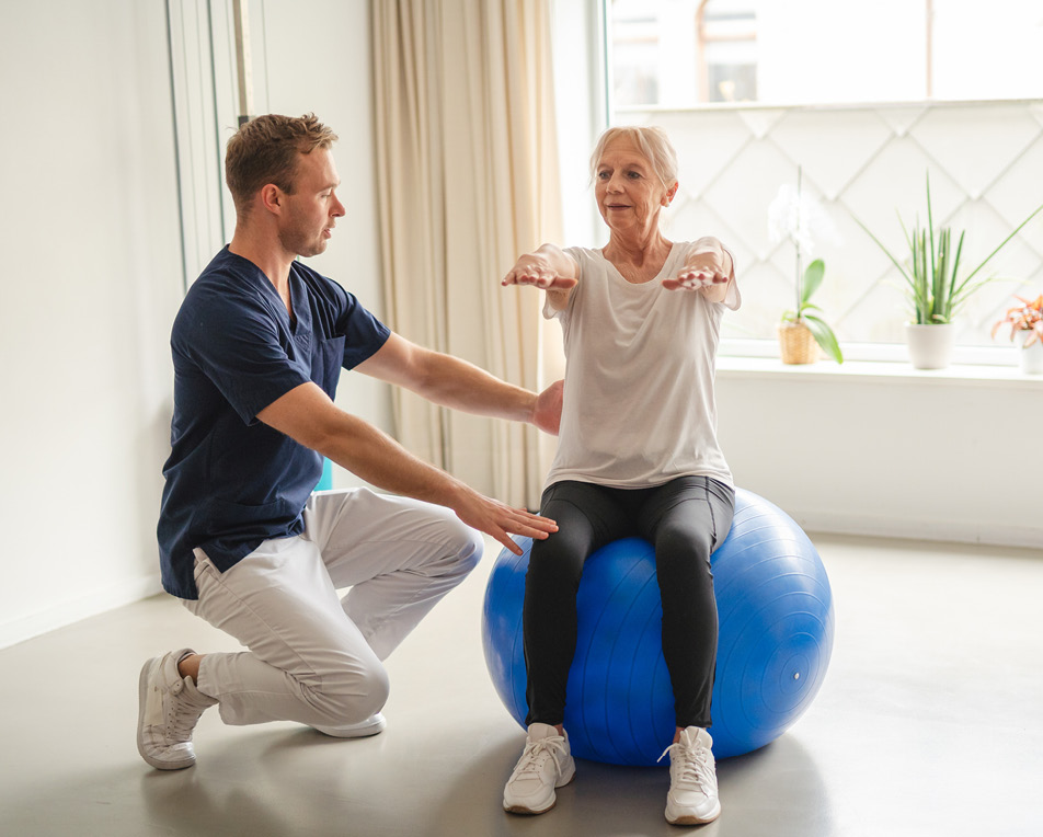 Vrouw zit op een blauwe yogabal en man helpt haar.