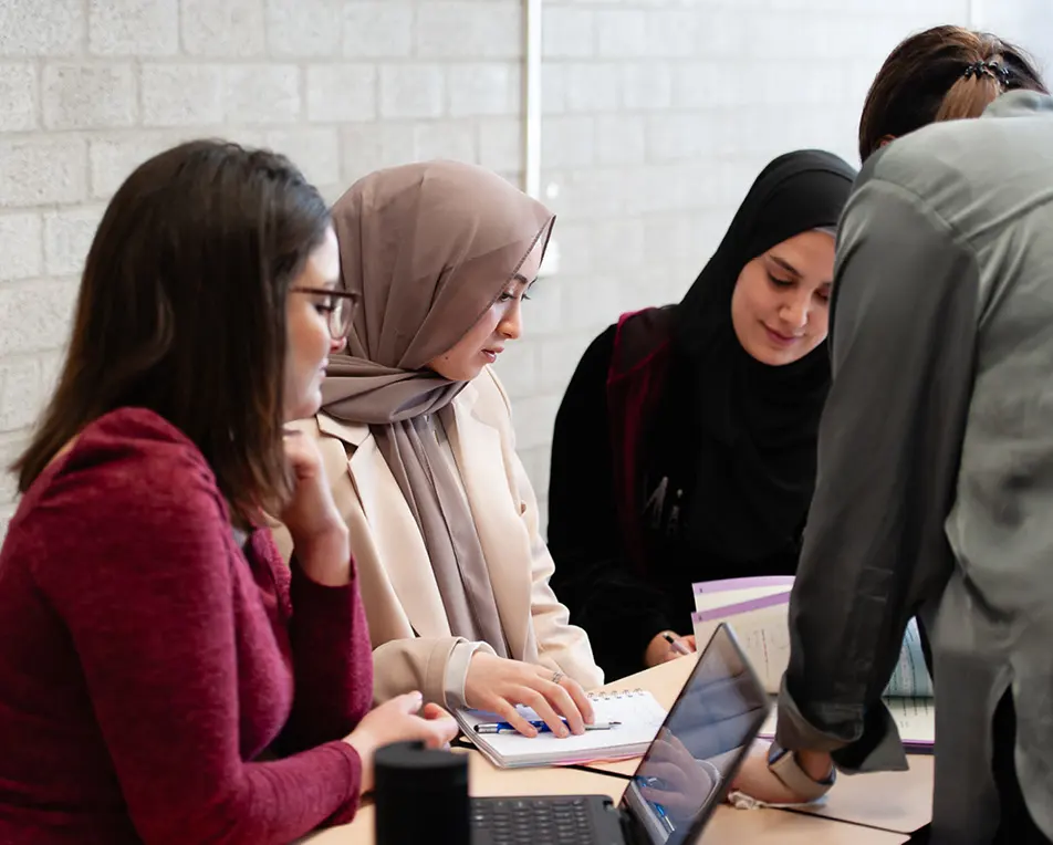 Vier personen rond een tafel met boeken en laptop, één persoon staat en legt iets uit