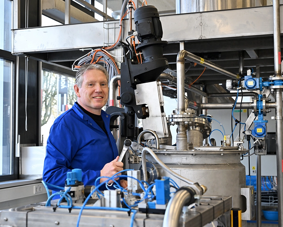 Een medewerker in een blauwe werkjas bedient industriële apparatuur in een technische installatie, omringd door metalen leidingen, sensoren en kabels.