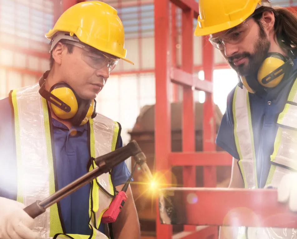 Twee personen met gele helmen en veiligheidshesjes werken in een industriële omgeving, waarbij één van hen een vonkend gereedschap gebruikt.
