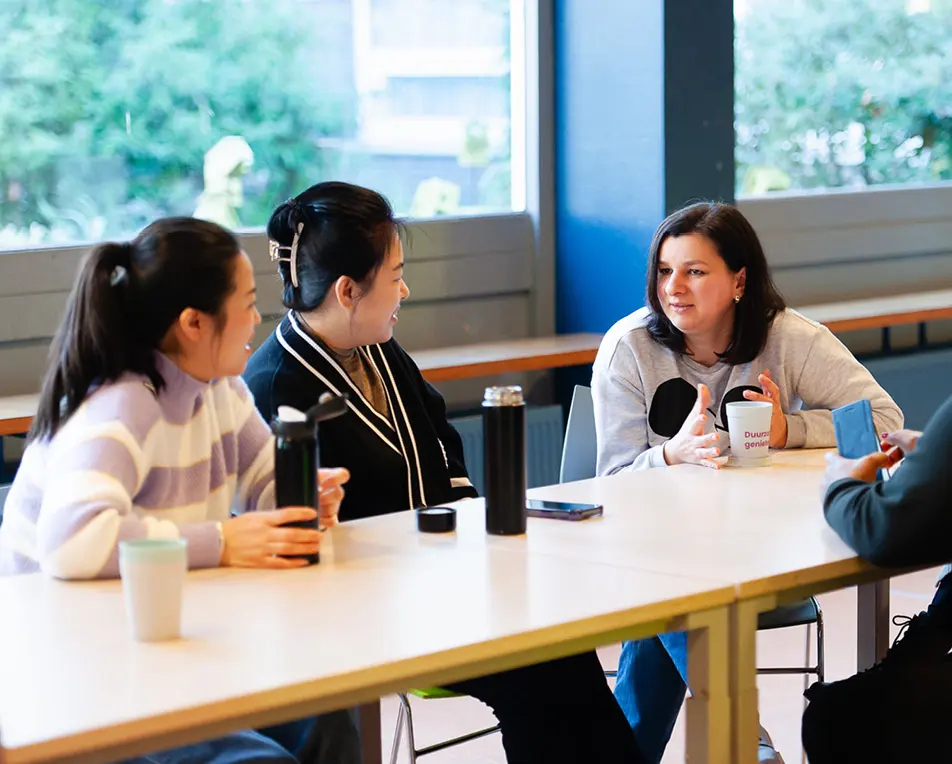 Drie personen zitten aan een bureau met boeken en papieren in een klas- of studieruimte.