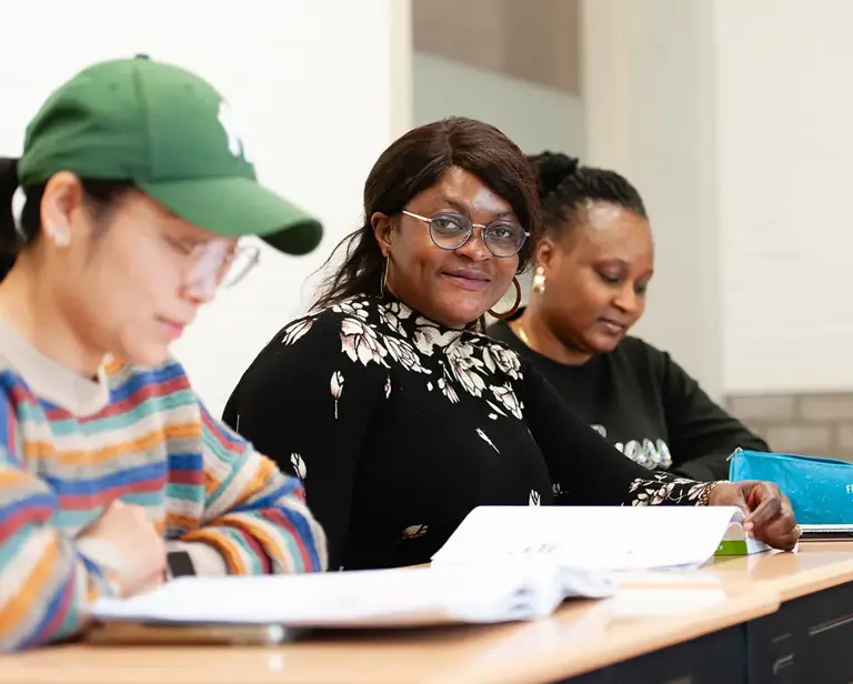 Vier personen zitten binnen rond een tafel in gesprek, met drankjes en een smartphone op tafel en grote ramen op de achtergrond.