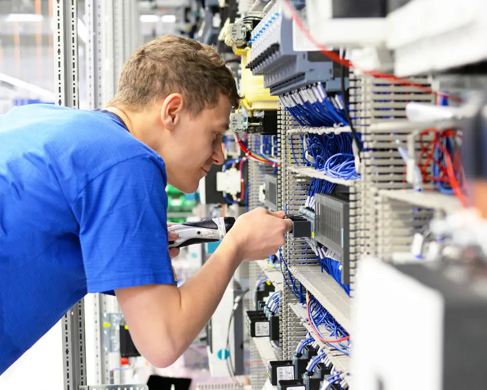 Een persoon in een blauw shirt werkt met gereedschap aan een groot elektrisch paneel vol bedrading in een technische omgeving.