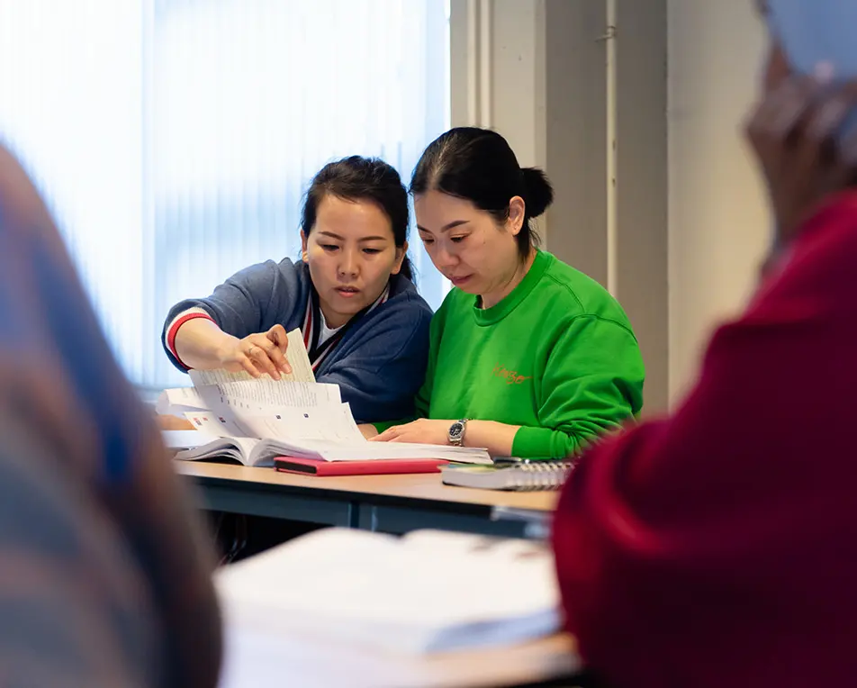 Twee personen zitten aan een tafel en bekijken samen een open boek, met extra boeken en notitieblokken eromheen.