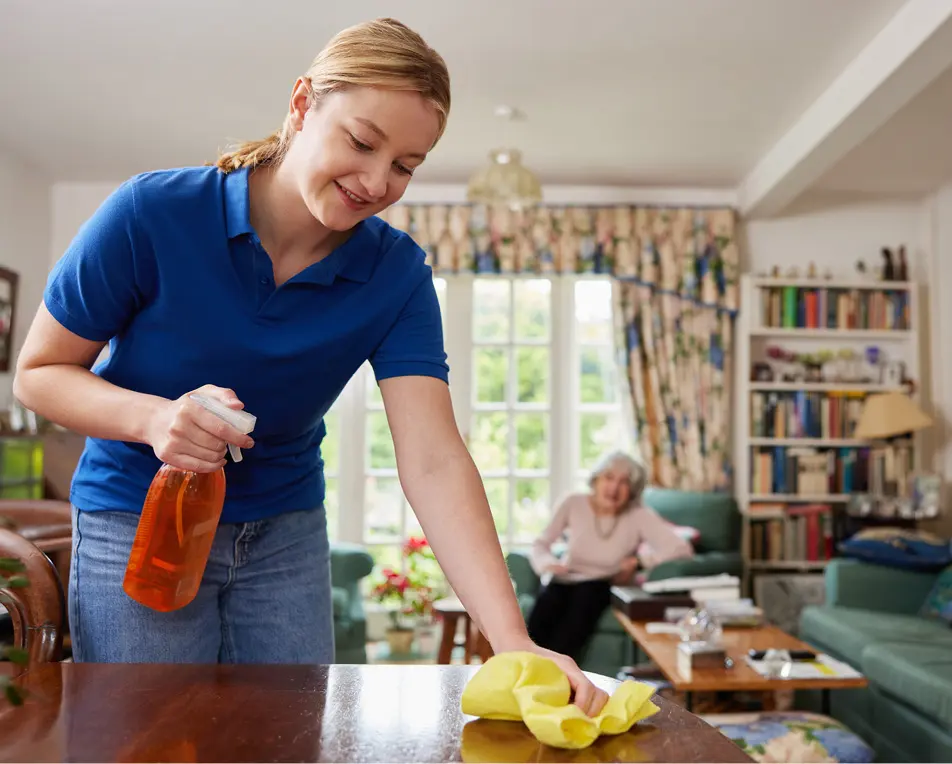 Huiskamer van een oudere vrouw, op de voorgrond is een vrouw in blauw t-shirt aan het poetsen.