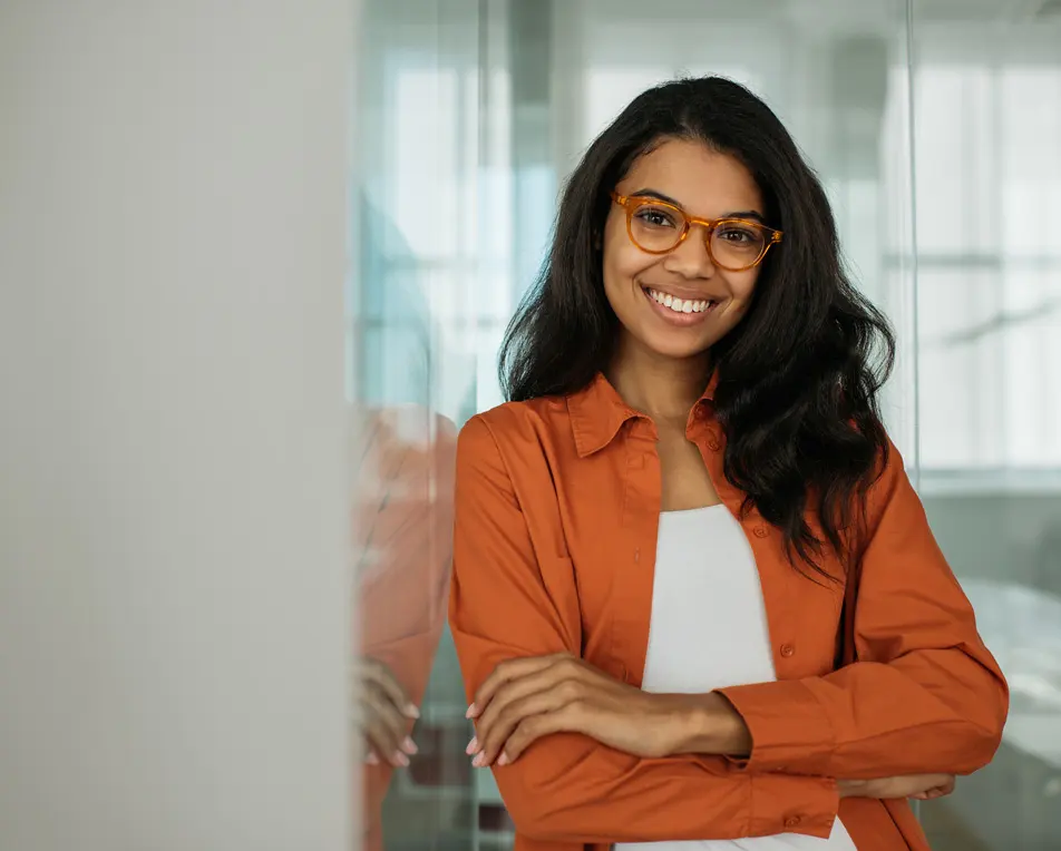 Vrouw met oranje jasje en bril leunt tegen de muur van een lichte ruimte aan.