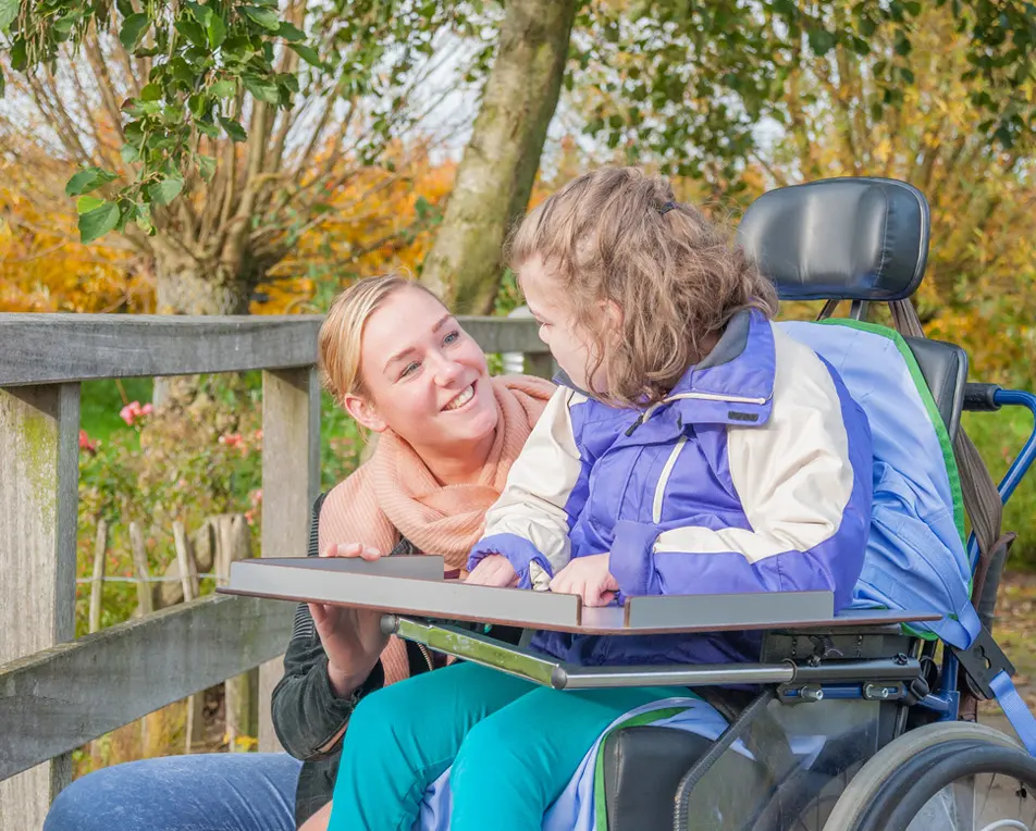 Blonde vrouw en meisje in rolstoel staan buiten in een park.