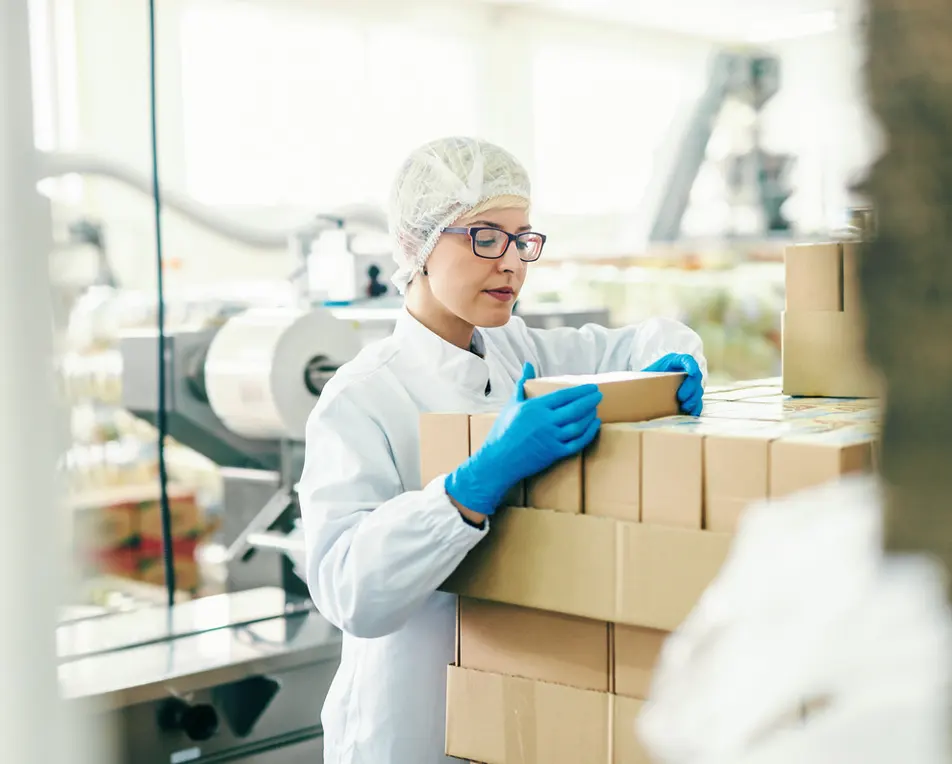 Fabrieksomgeving met vrouw in witte outfit die een doos vast heeft.
